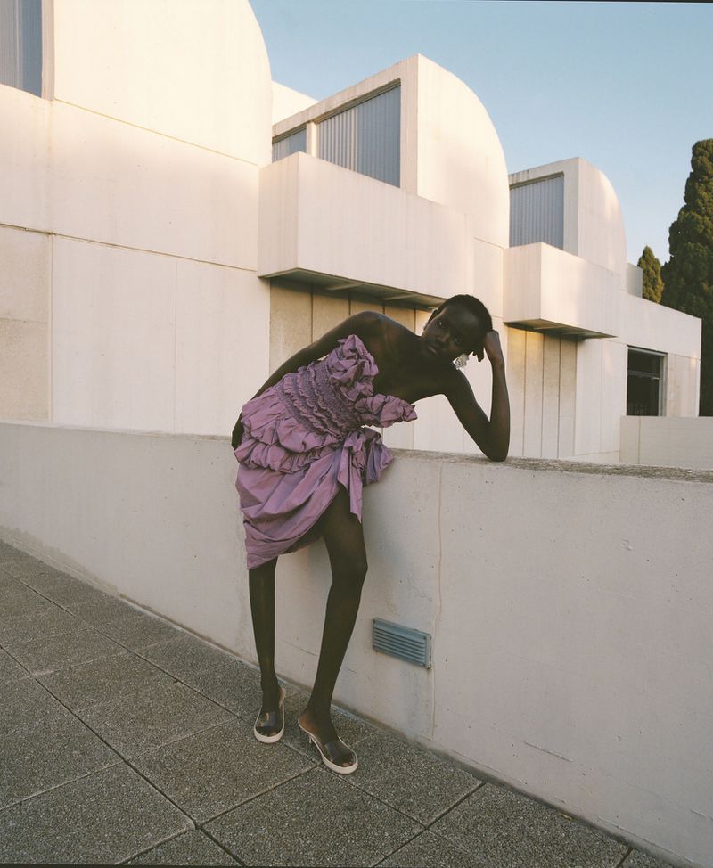 Model Yar Aguer in a purple ruffled dress leaning against a modern white concrete building exterior for Vogue Espana.