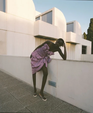 Model Yar Aguer in a purple ruffled dress leaning against a modern white concrete building exterior for Vogue Espana.