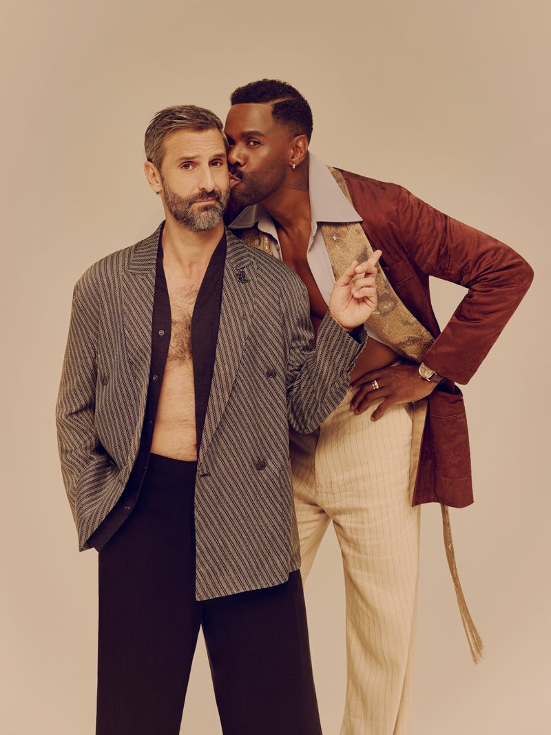 Two men posing against a neutral backdrop, one in a striped double-breasted blazer and another in a burgundy coat kissing his cheek