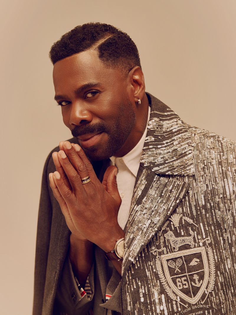 Portrait of Colman Domingo clasping hands together, wearing an embellished grey jacket and white shirt against a neutral background.