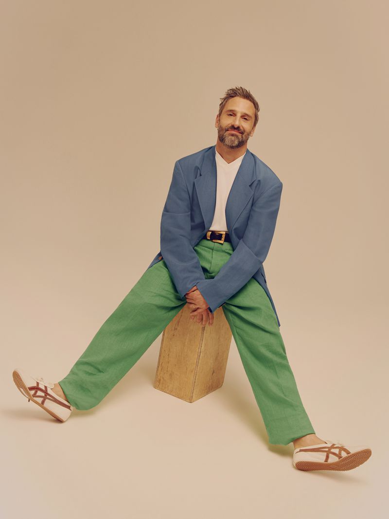 Portrait of Marco Calvani seated on a wooden block wearing a blue blazer and green trousers against a neutral background