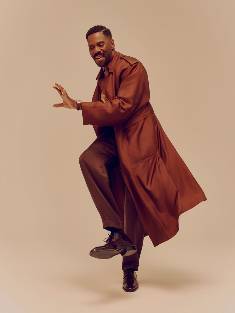 Actor Colman Domingo striking a joyful pose in a brown trench coat against a warm neutral studio backdrop for Out Magazine.