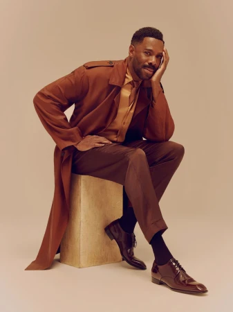 Colman Domingo seated on a wooden block wearing a brown coat and trousers against a neutral background