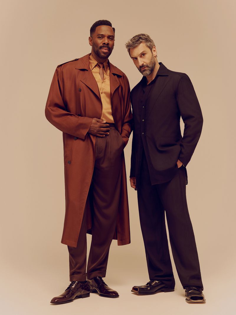 Colman Domingo in a brown trench coat and Marco Calvani in a black suit standing against a seamless neutral background.