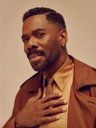 Colman Domingo wearing a brown coat and tan collared shirt with a tie against a warm neutral background.