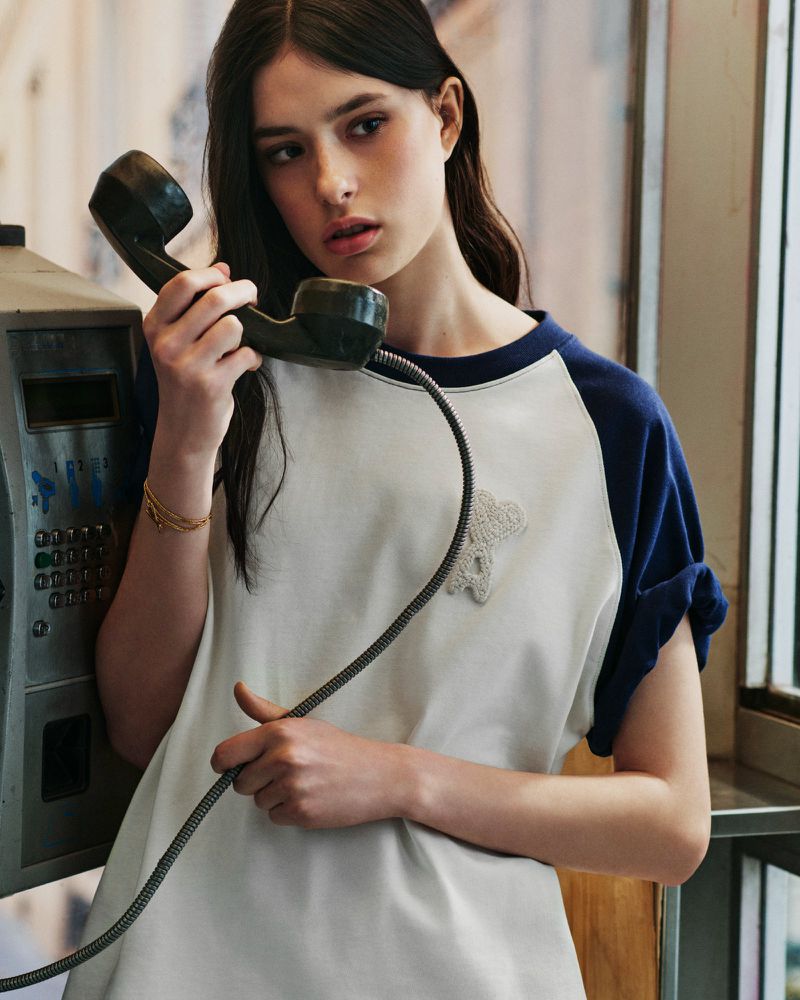 Young woman holding a payphone receiver wearing a white t shirt with blue sleeves for Ami