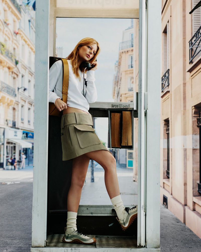 A woman in a white long-sleeve top and olive green utility skirt stands inside a street telephone booth holding the receiver to her ear.
