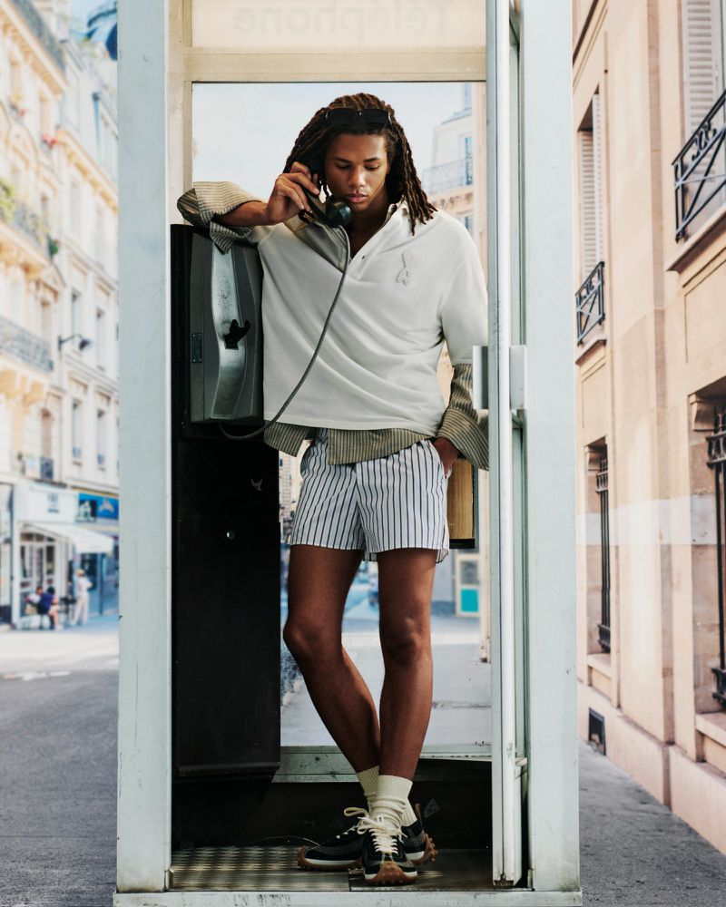 Man with dreadlocks wearing striped shorts and white polo shirt standing inside a glass telephone booth on a city street.