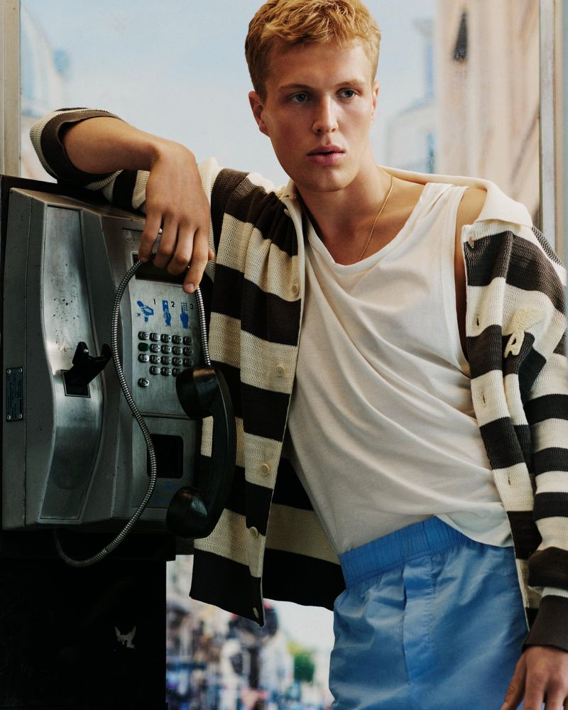 Blonde male model leaning against a vintage payphone wearing a striped cardigan, white tank top, and blue trousers.