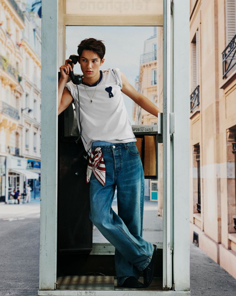 Man stands in a telephone booth on a city street, wearing a white t-shirt and jeans, holding the phone to his ear in daytime lighting.
