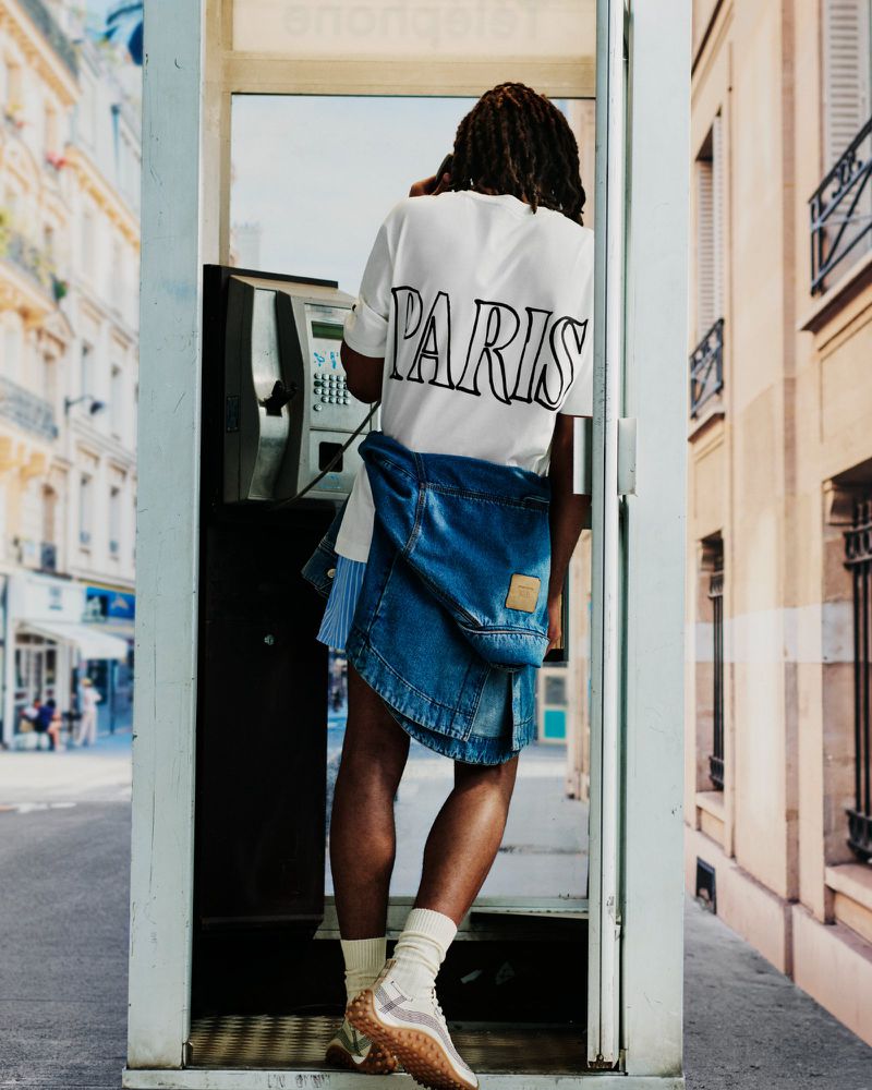 Man with dreadlocks wearing a white Paris t-shirt and denim jacket tied around his waist inside a telephone booth on a city street.