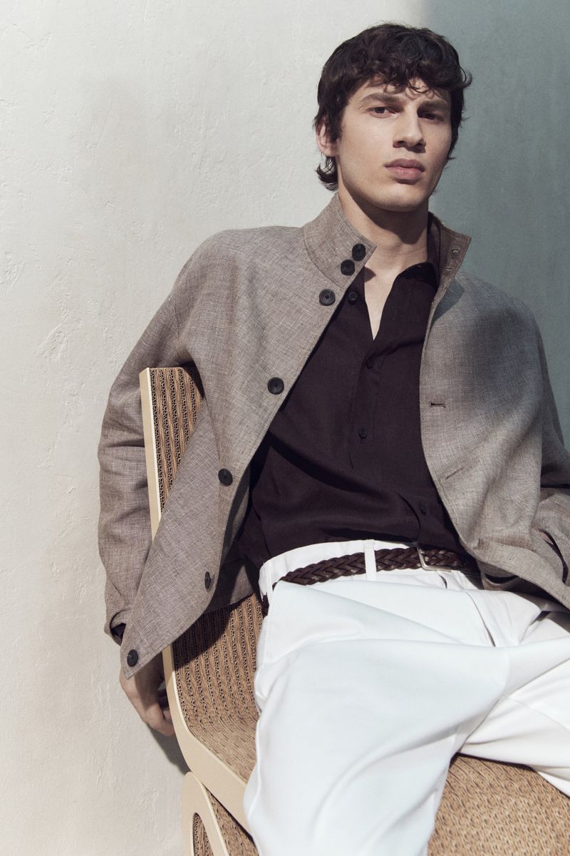 A male model wearing a grey jacket and white trousers seated on a chair against a textured wall for Massimo Dutti