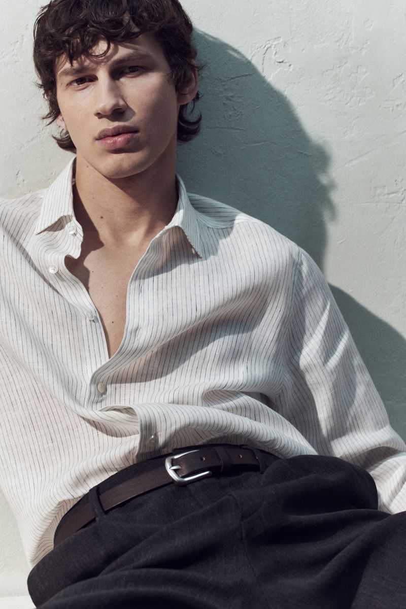 Close-up portrait of a male model wearing a striped button-down shirt and dark trousers against a light textured wall.