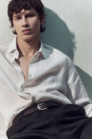 Close-up portrait of a male model wearing a striped button-down shirt and dark trousers against a light textured wall.