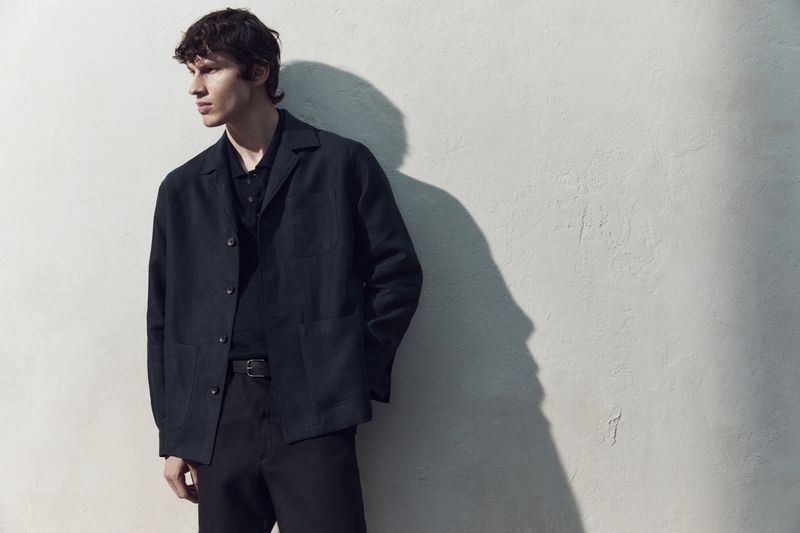 A man in a dark jacket and polo shirt stands against a pale textured wall, casting a sharp shadow.