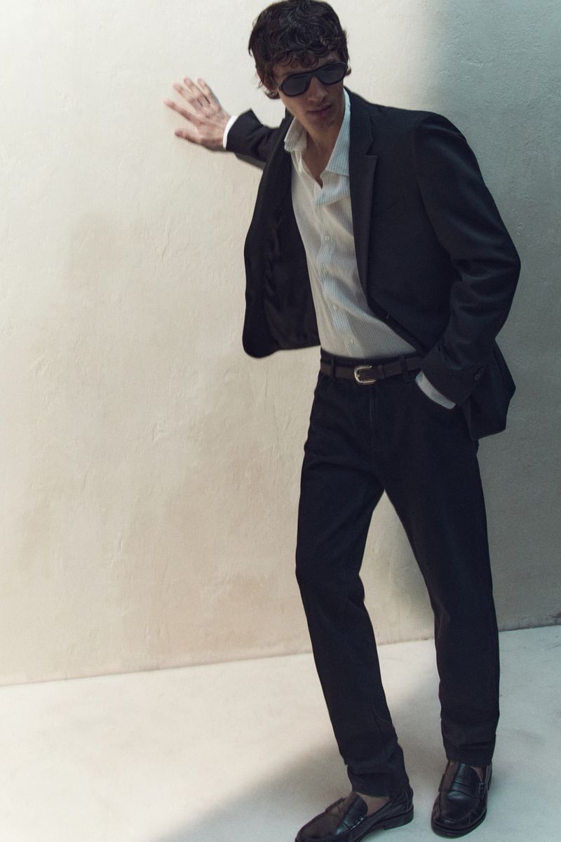 Man wearing a black suit jacket, white shirt, and dark trousers leaning against a neutral studio wall.