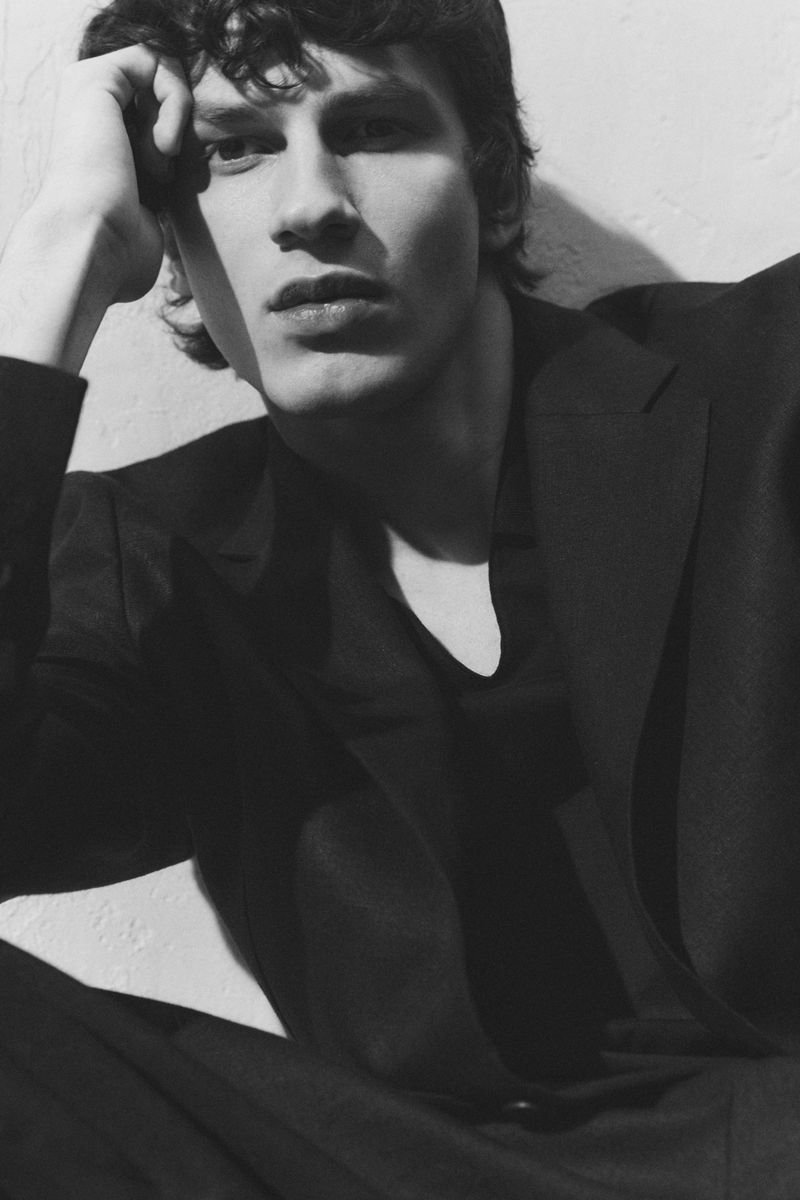 Monochrome portrait of a male model wearing a dark suit jacket and resting his hand near his head against a plain wall