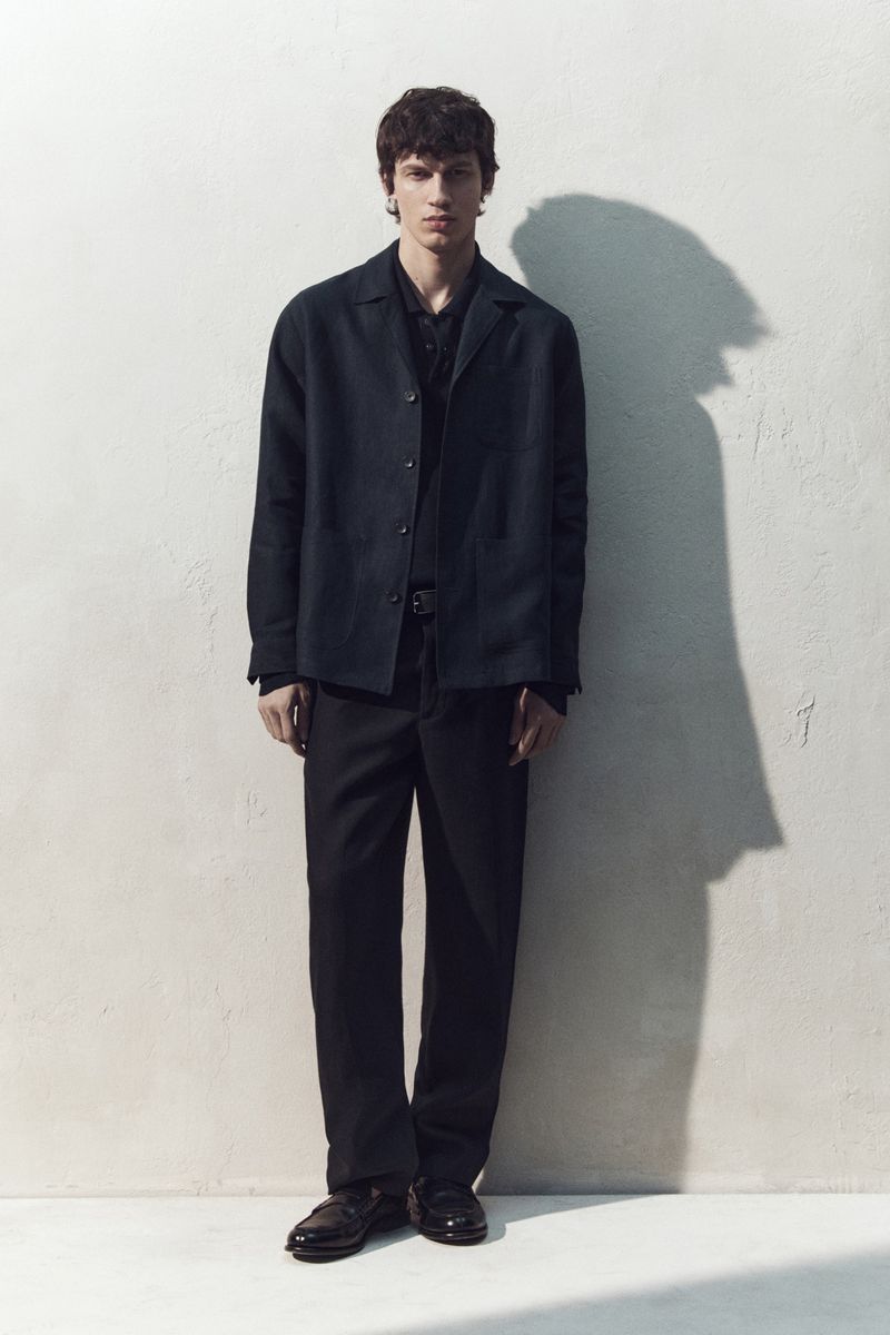 Full-length portrait of a male model wearing a dark jacket and trousers against a textured white wall casting a sharp shadow