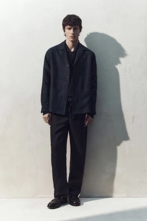 Full-length portrait of a male model wearing a dark jacket and trousers against a textured white wall casting a sharp shadow