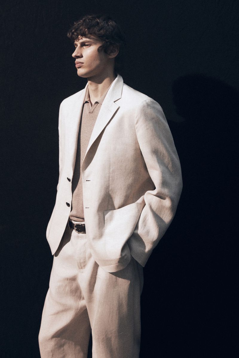 Male model in a cream linen suit and polo shirt standing against a dark background for Massimo Dutti summer looks