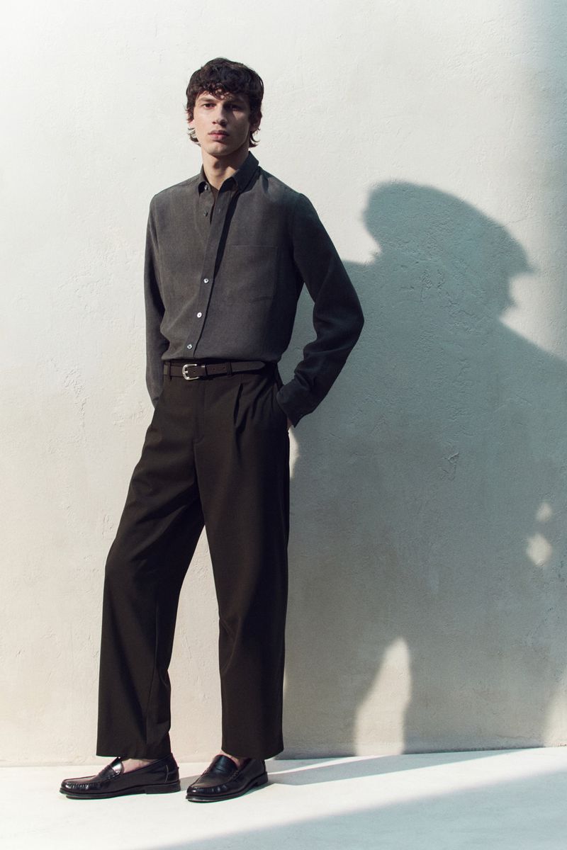A young adult male poses in a grey shirt and black trousers against a textured wall, bathed in natural light and shadows.