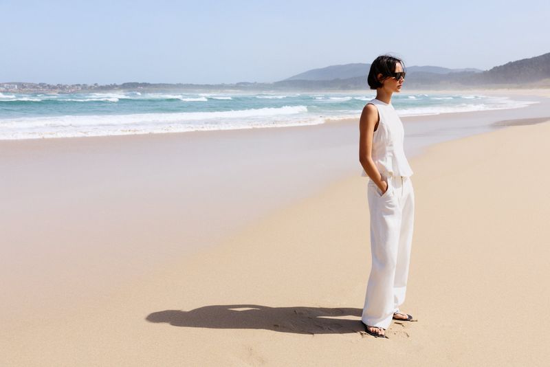 Model wearing white sleeveless top and matching trousers standing on a sandy beach looking toward ocean waves