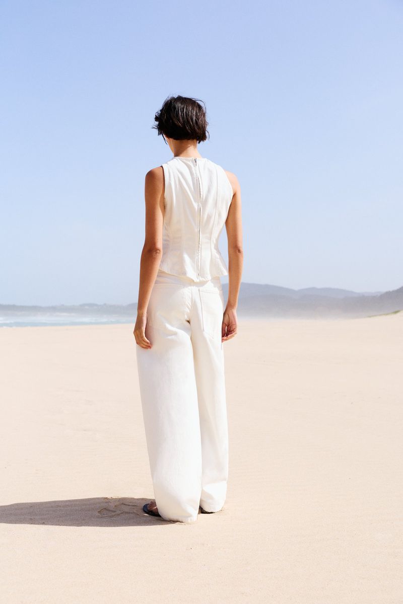 Model seen from behind wearing a white sleeveless top and matching trousers standing on a sunny sandy beach