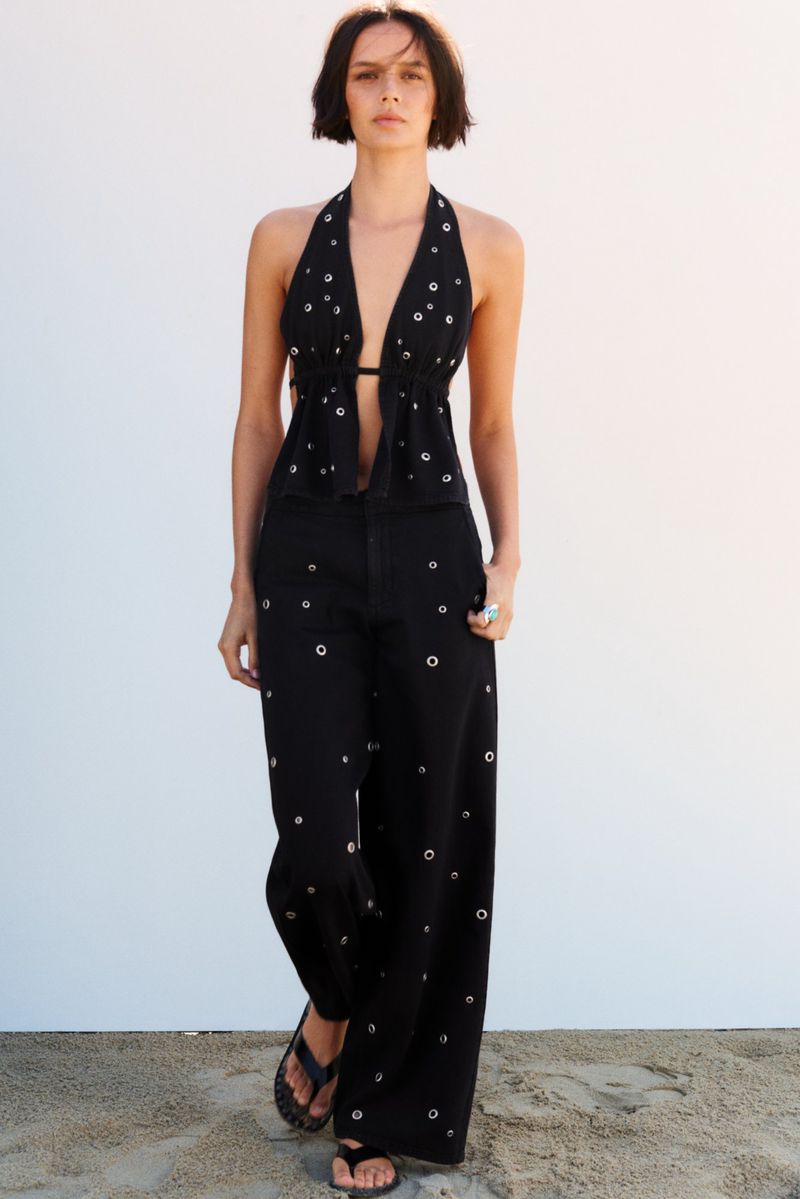 A woman models a black halter top and wide-leg pants with grommet accents, complemented by dark sandals on a sunny backdrop.