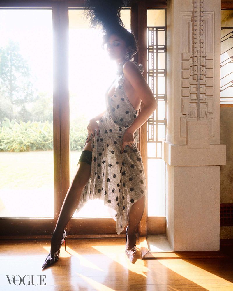 Zendaya wearing a polka dot dress and feathered headpiece standing by a large window for Vogue Brasil