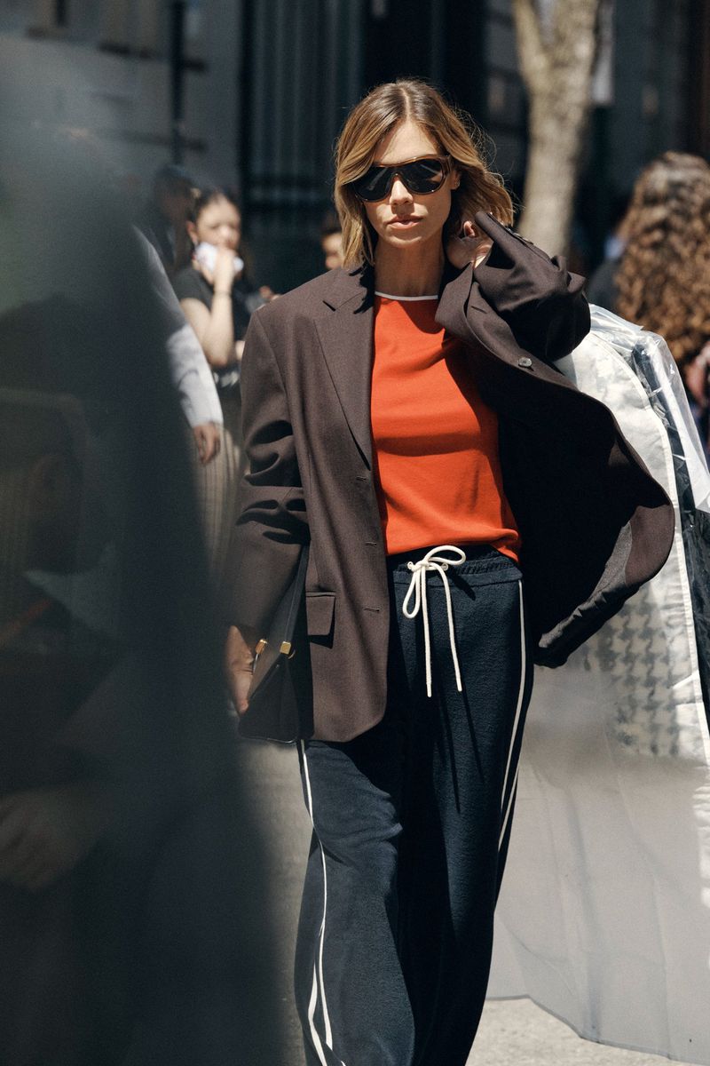 Model wearing dark sunglasses, brown blazer, orange top, and dark trousers outdoors in an urban setting