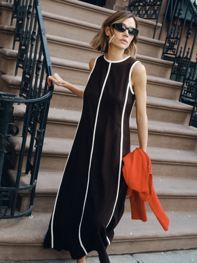 Model Joanna Halpin in dark sunglasses and a black maxi dress standing on brown stone steps holding a bright red sweater.