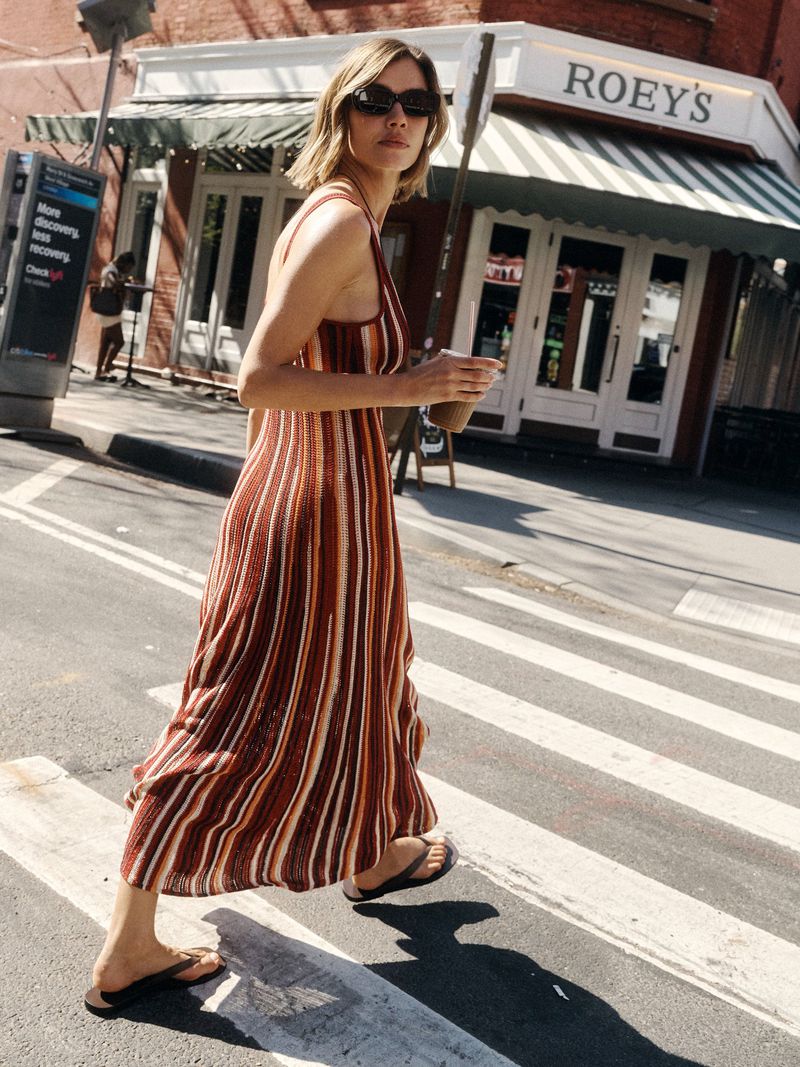Woman in a striped brown knit dress walking across a city street carrying a iced drink past a storefront.