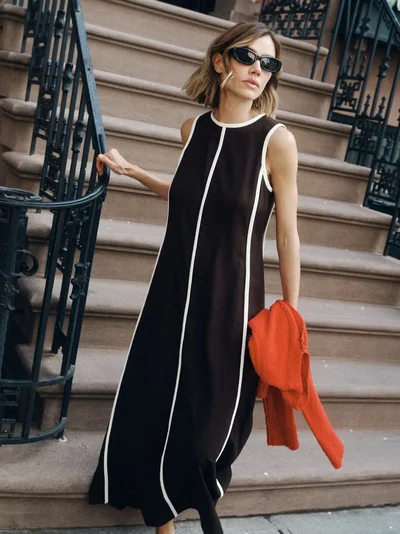 Model Joanna Halpin in dark sunglasses and a black maxi dress standing on brown stone steps holding a bright red sweater.