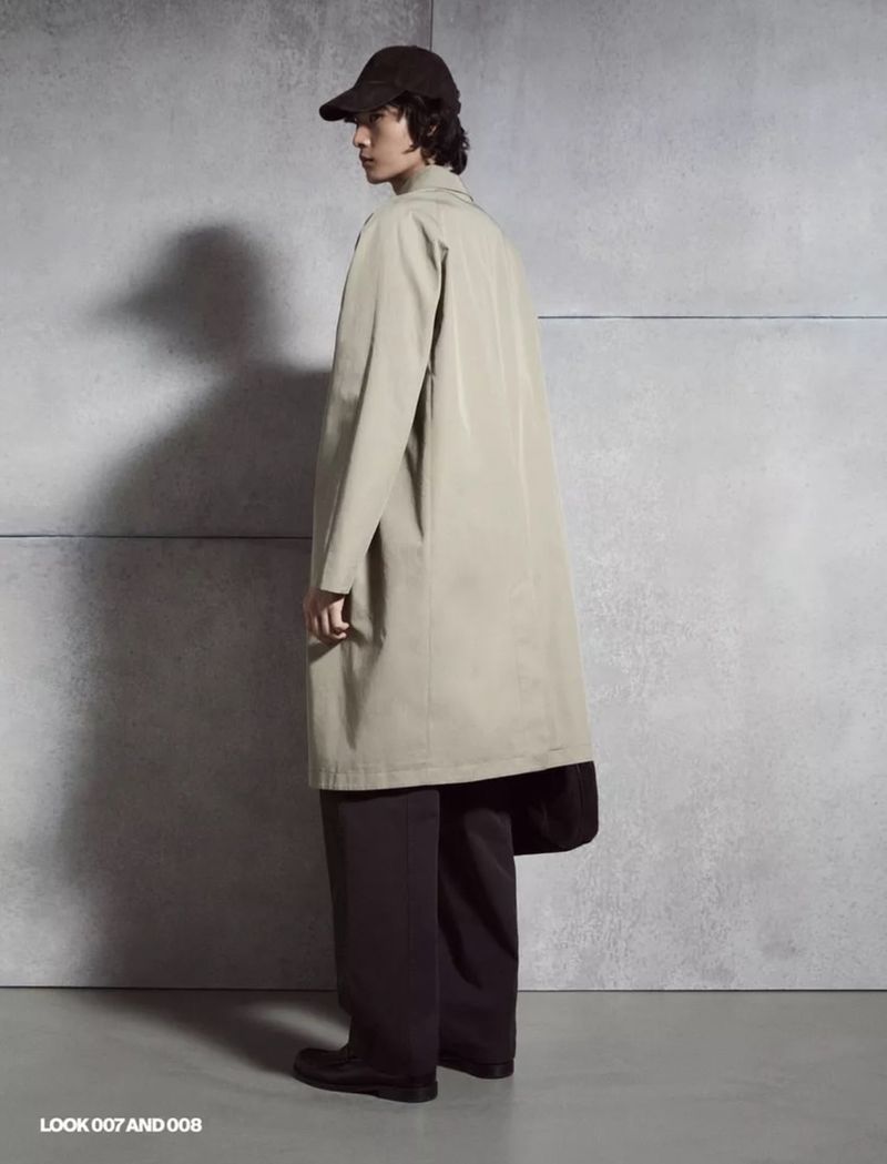 A person is seen in a fashion shot, facing away, wearing a long light-brown trench coat, dark cap, and dark wide-leg pants, against a neutral wall.