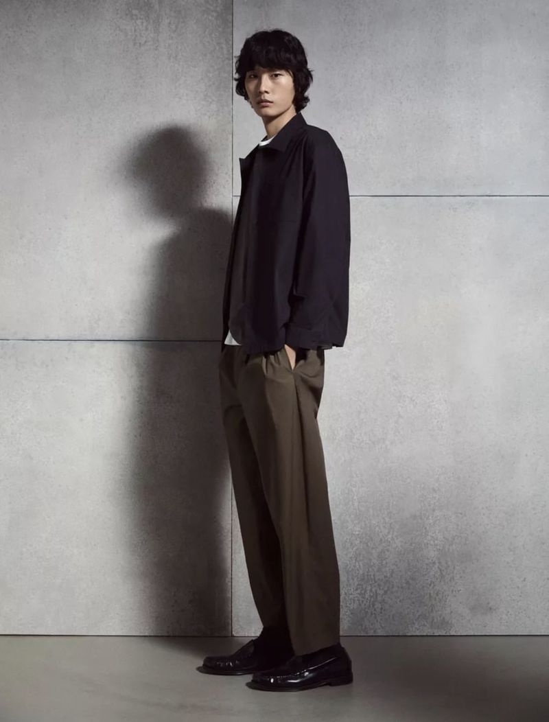 Model standing against a grey concrete wall wearing a dark jacket, brown trousers, and loafers for Massimo Dutti