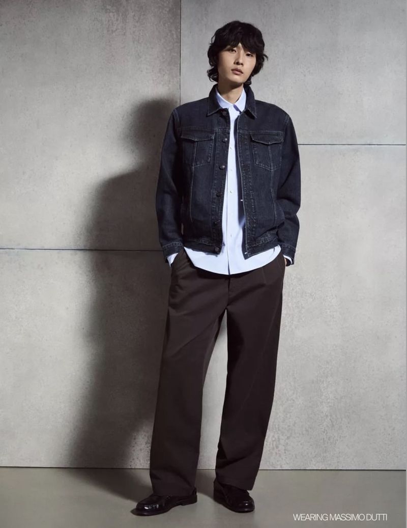 Model in dark denim jacket and brown trousers against a concrete textured studio background