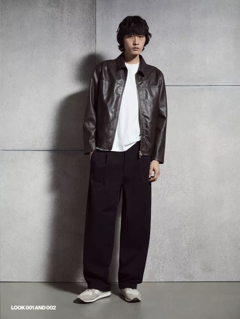 Model wearing a brown leather jacket, white t-shirt, and dark trousers standing against a textured concrete wall.