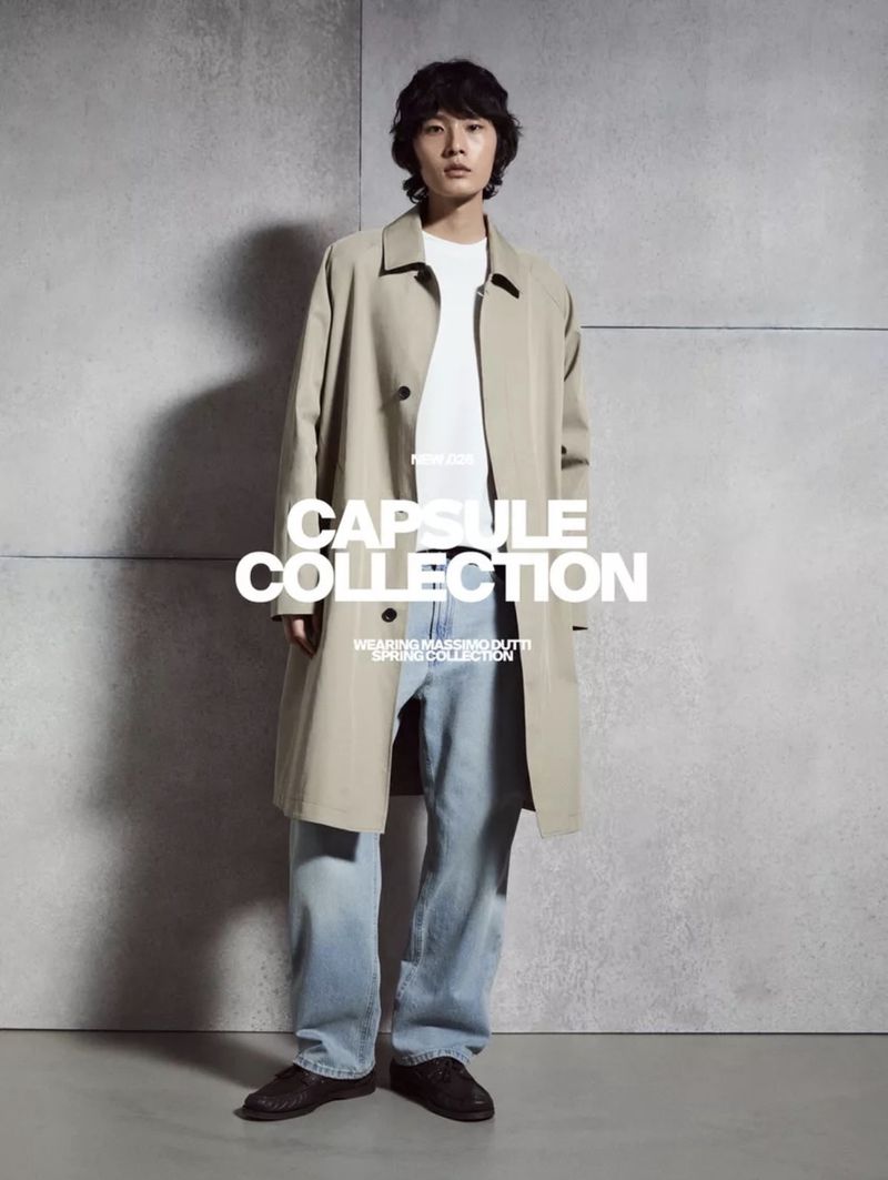 A model wearing a beige trench coat, white t-shirt, and blue jeans standing against a grey concrete wall.
