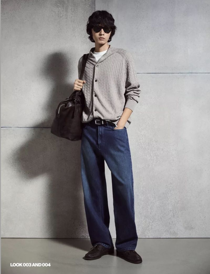 Model in dark sunglasses, beige knit cardigan, and blue denim jeans holding a dark leather duffel bag against a concrete texture wall.