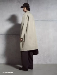 Model wearing a long beige trench coat and dark baseball cap standing against a grey concrete wall in profile.