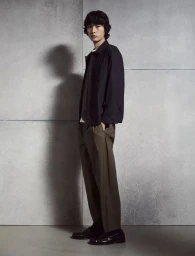 Model standing against a grey concrete wall wearing a dark jacket, brown trousers, and loafers for Massimo Dutti