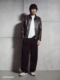 Model wearing a brown leather jacket, white t-shirt, and dark trousers standing against a textured concrete wall.