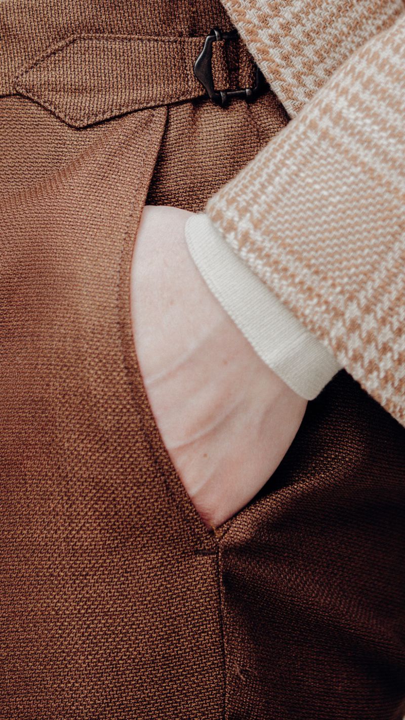 Close-up detail of a man's hand in his pocket wearing brown tailored trousers and a patterned jacket