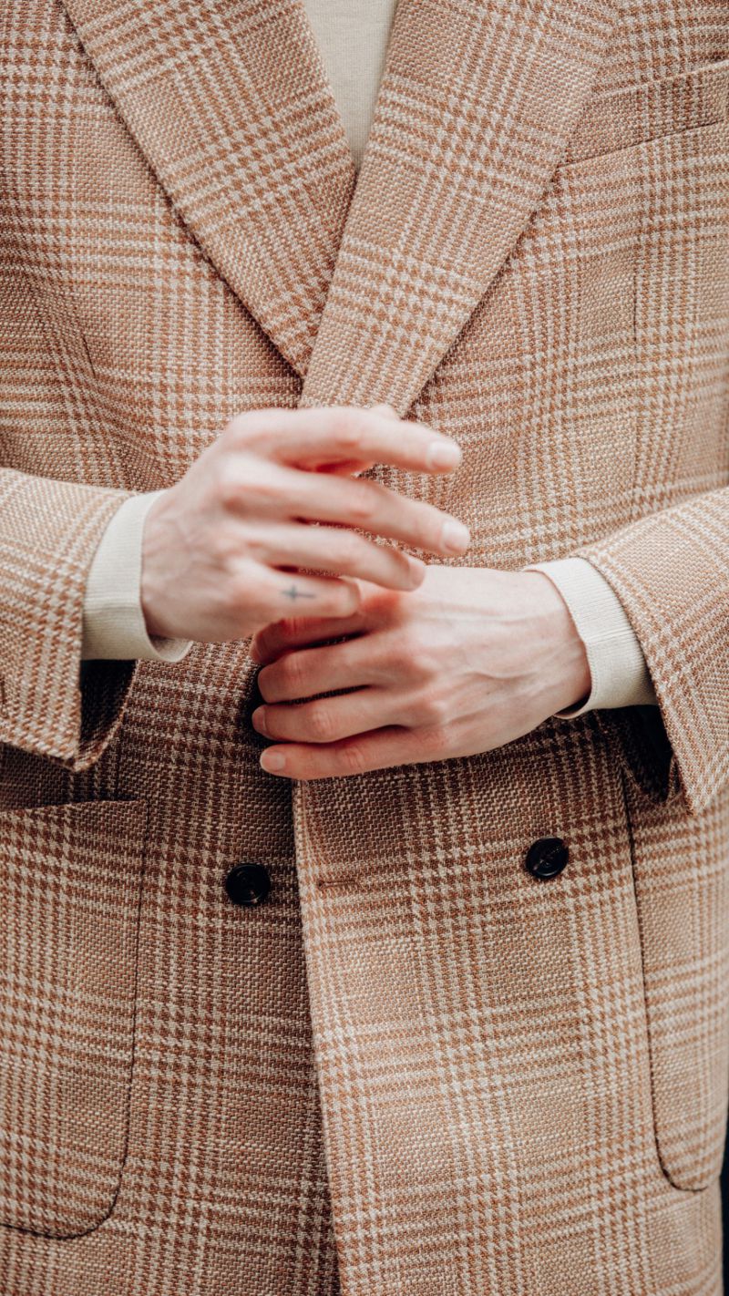 Close-up of a man wearing a brown plaid tailored blazer with black buttons over a neutral sweater, adjusting his cuffs.