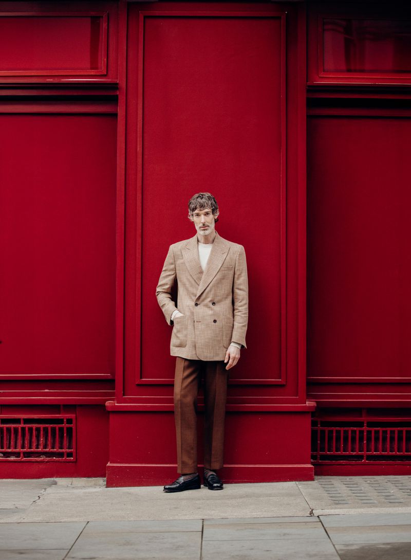 Stylish adult man in double-breasted blazer and brown trousers poses against a red wall in an urban setting. Fashion editorial shot.
