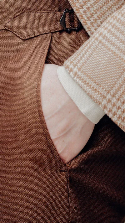 Close-up detail of a man's hand in his pocket wearing brown tailored trousers and a patterned jacket