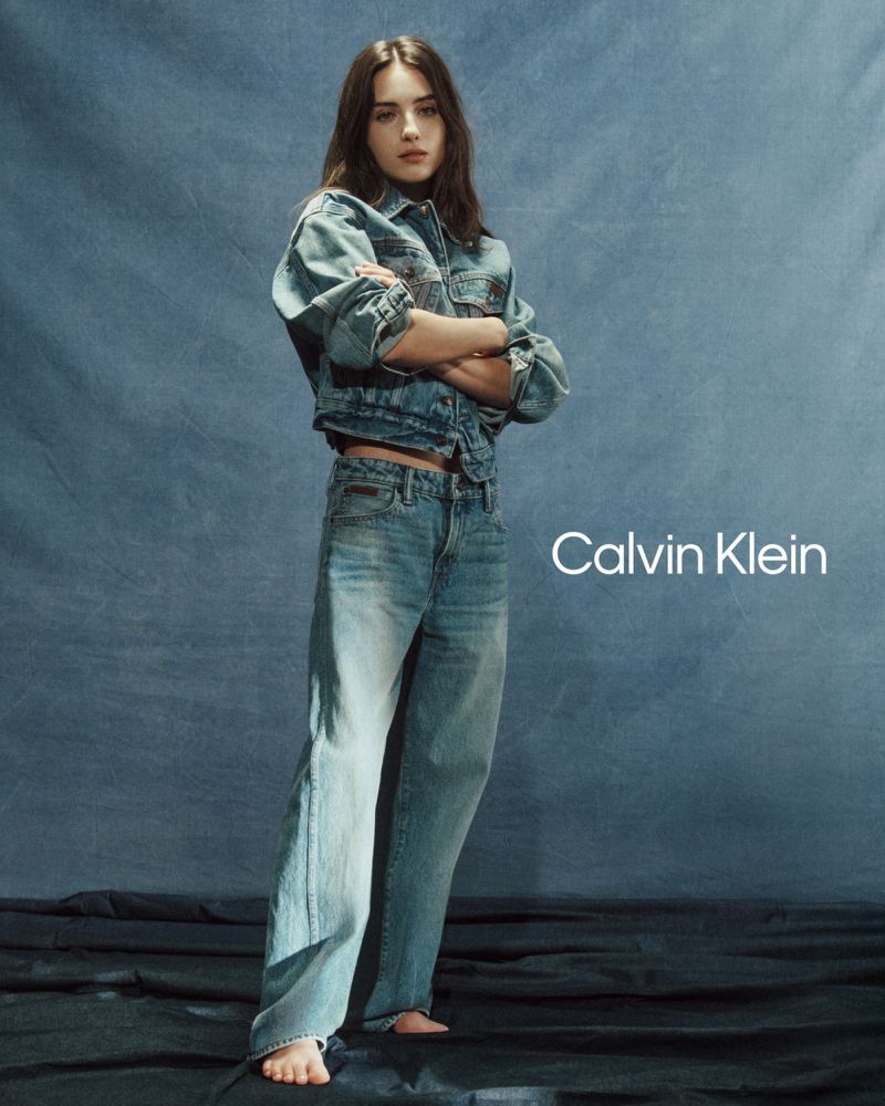 A young woman in a denim jacket and jeans poses with arms crossed in a studio setting for a Calvin Klein fashion shot.
