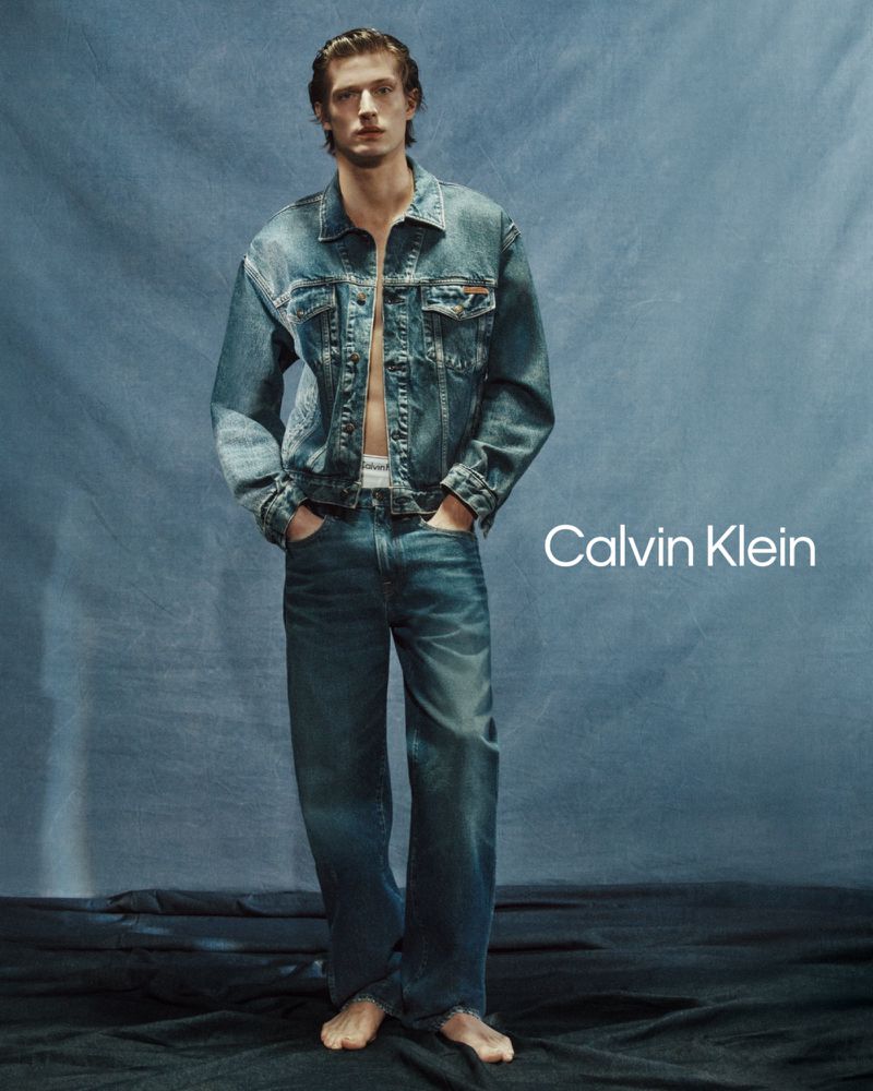 Male model poses in a denim jacket and jeans against a blue backdrop, showcasing the Calvin Klein brand logo.