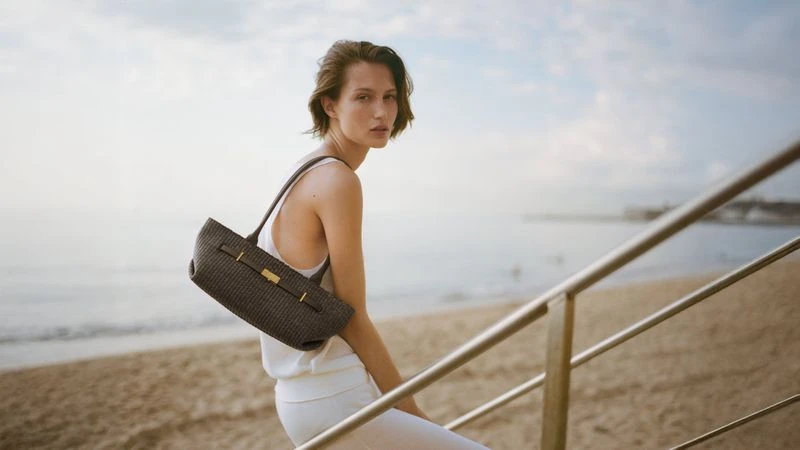 Model Vika Evseeva wearing white apparel and holding a dark woven handbag by the beach for DeMellier London