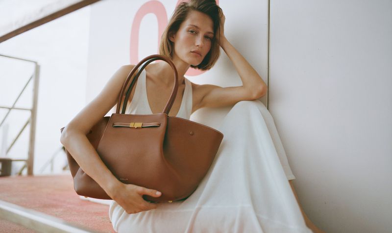 Model wearing white top holding large brown leather handbag against light wall for DeMellier London